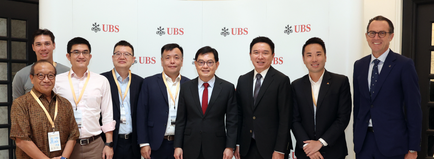 Nine people stand in front of UBS logos wearing business attire and lanyards.